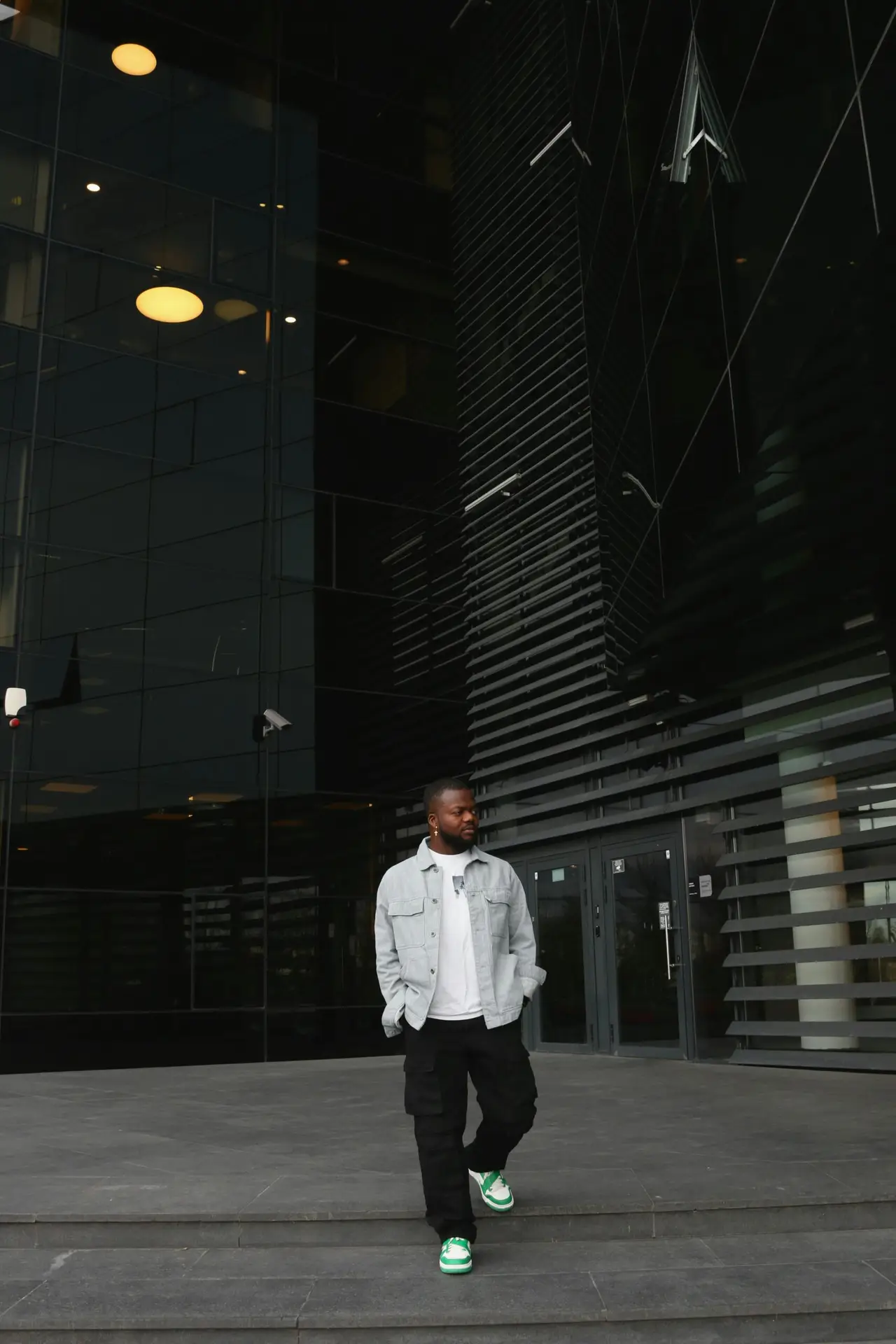 A stylish man walks in front of a modern glass building, showcasing urban fashion and architecture.