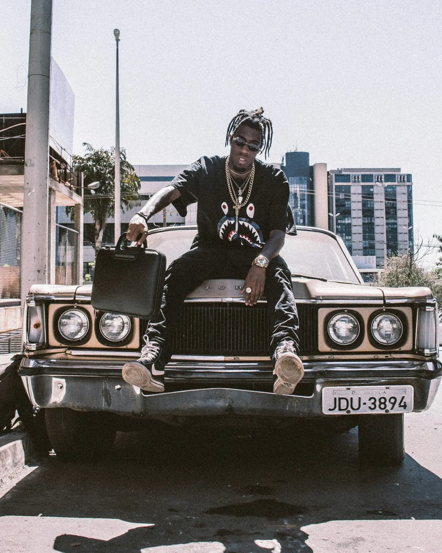 A fashionable young man poses on a vintage car in an urban cityscape, exuding streetwear style.