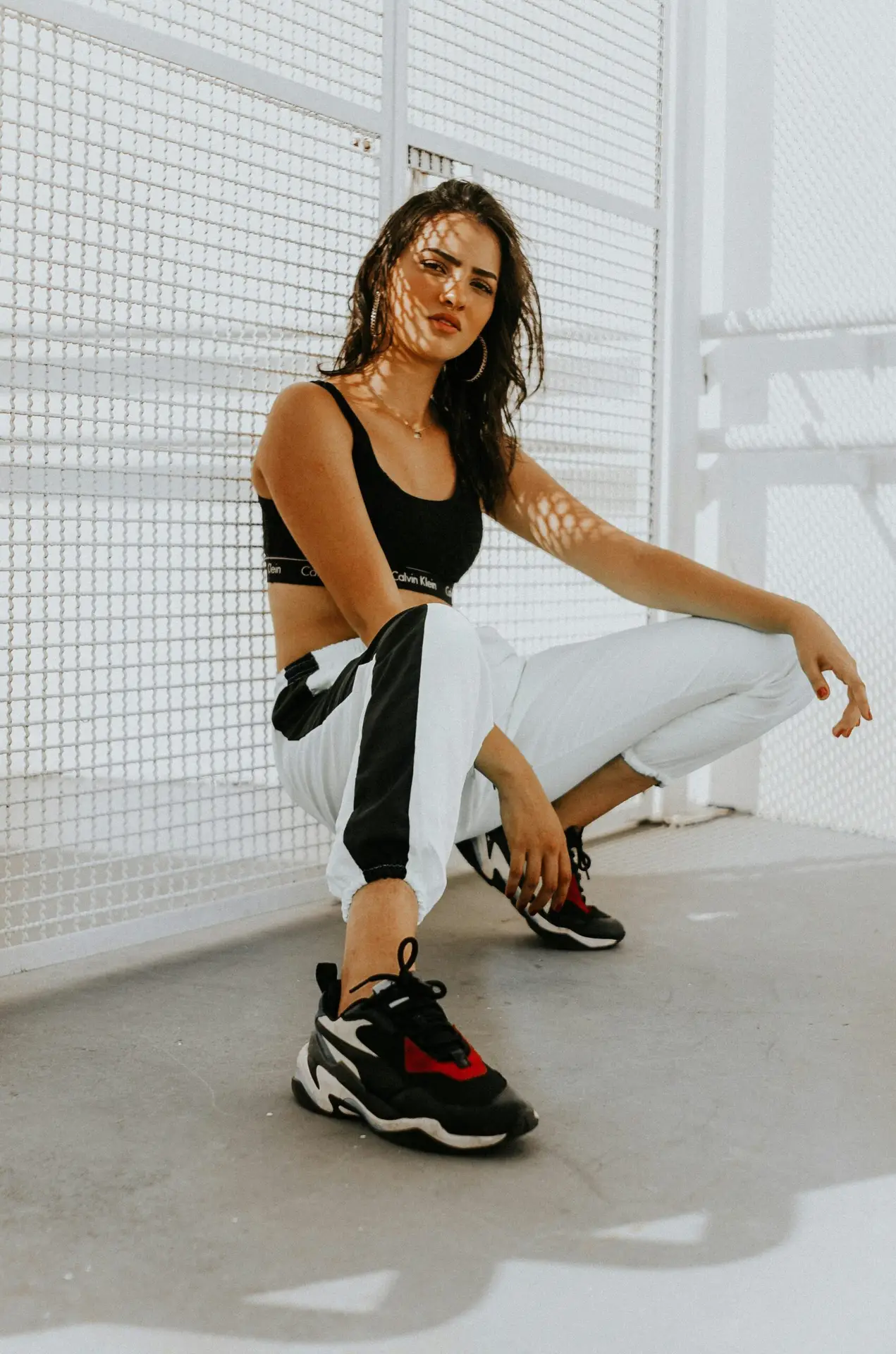 Confident woman in urban attire, featuring trendy sneakers, crouching in sunlight.