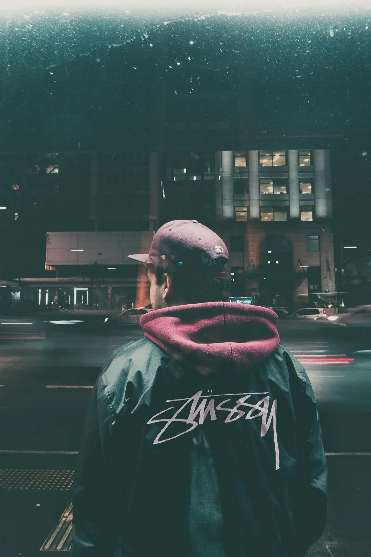 Man standing in city at night wearing a stylish jacket with illuminated buildings and passing traffic.